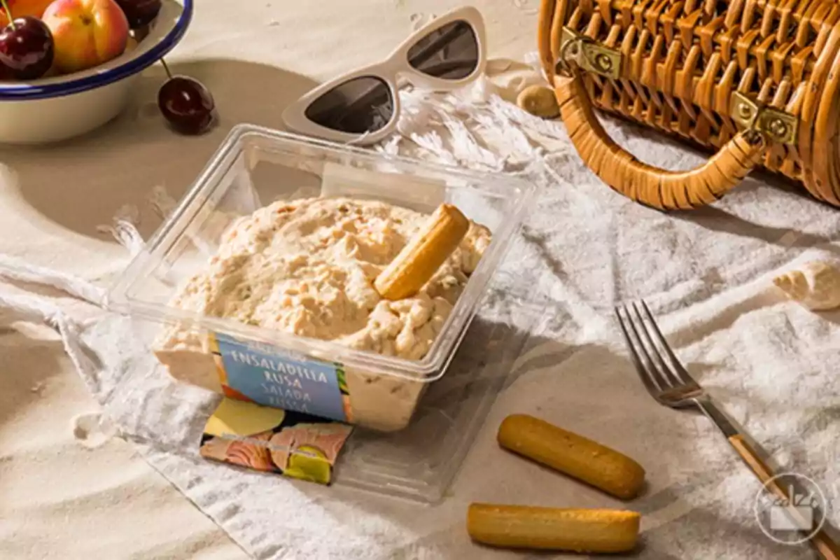 Ensaladilla rusa en un envase de plástico acompañada de palitos de pan sobre un mantel junto a un tenedor, unas gafas de sol, una cesta de mimbre y un bol de frutas Ensaladilla rusa en un envase de plástico acompañada de palitos de pan sobre un mantel junto a un tenedor, unas gafas de sol, una cesta de mimbre y un bol de frutas