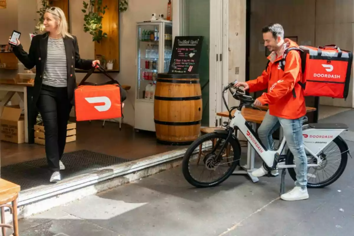 A person leaves an establishment with a delivery bag while another person on a bicycle waits outside, both wearing gear from a delivery company. A person leaves an establishment with a delivery bag while another person on a bicycle waits outside, both wearing gear from a delivery company.