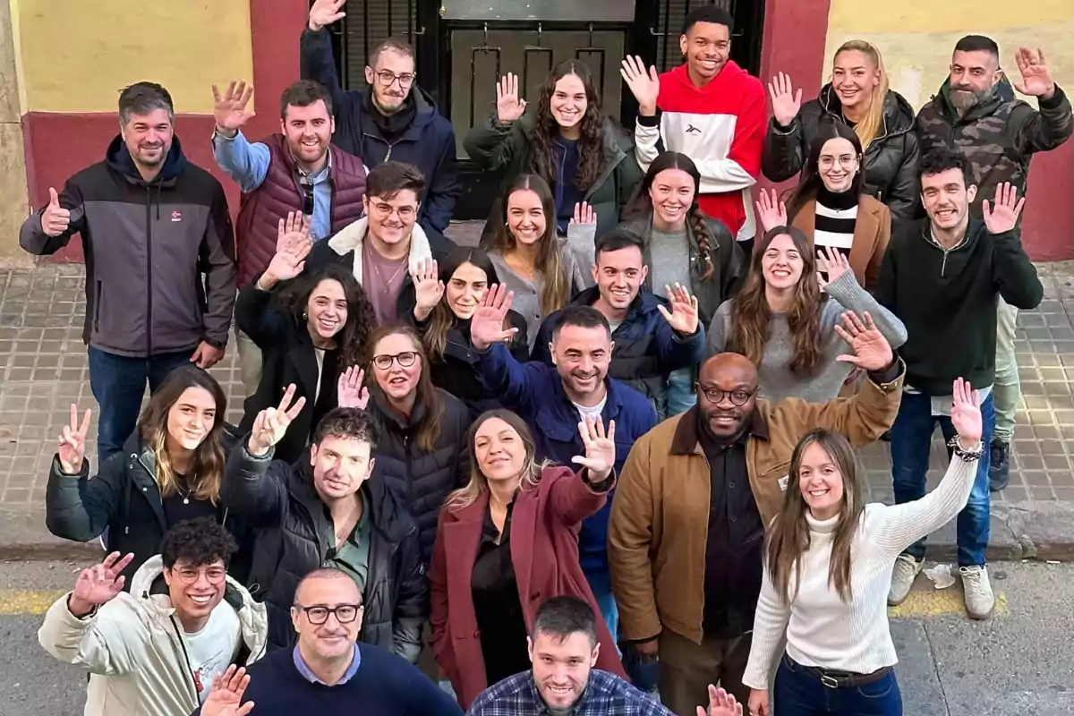 Un grupo de personas sonrientes posando al aire libre mientras levantan las manos en señal de saludo.