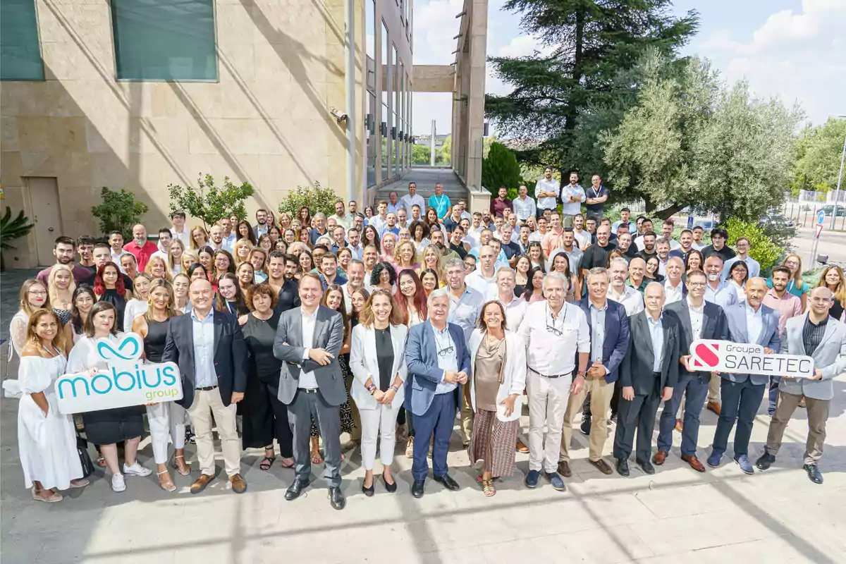 Un grupo grande de personas posando para una foto al aire libre, algunas sosteniendo carteles con los nombres "mobius group" y "Groupe SARETEC".