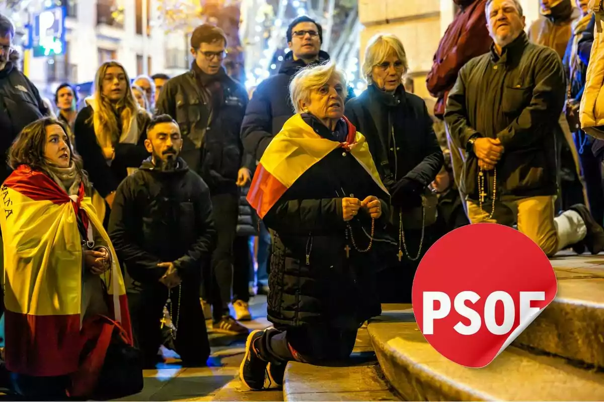 Un grupo de personas se encuentra arrodillado en la calle, algunas de ellas envueltas en banderas de España y sosteniendo rosarios, mientras otras observan de pie.