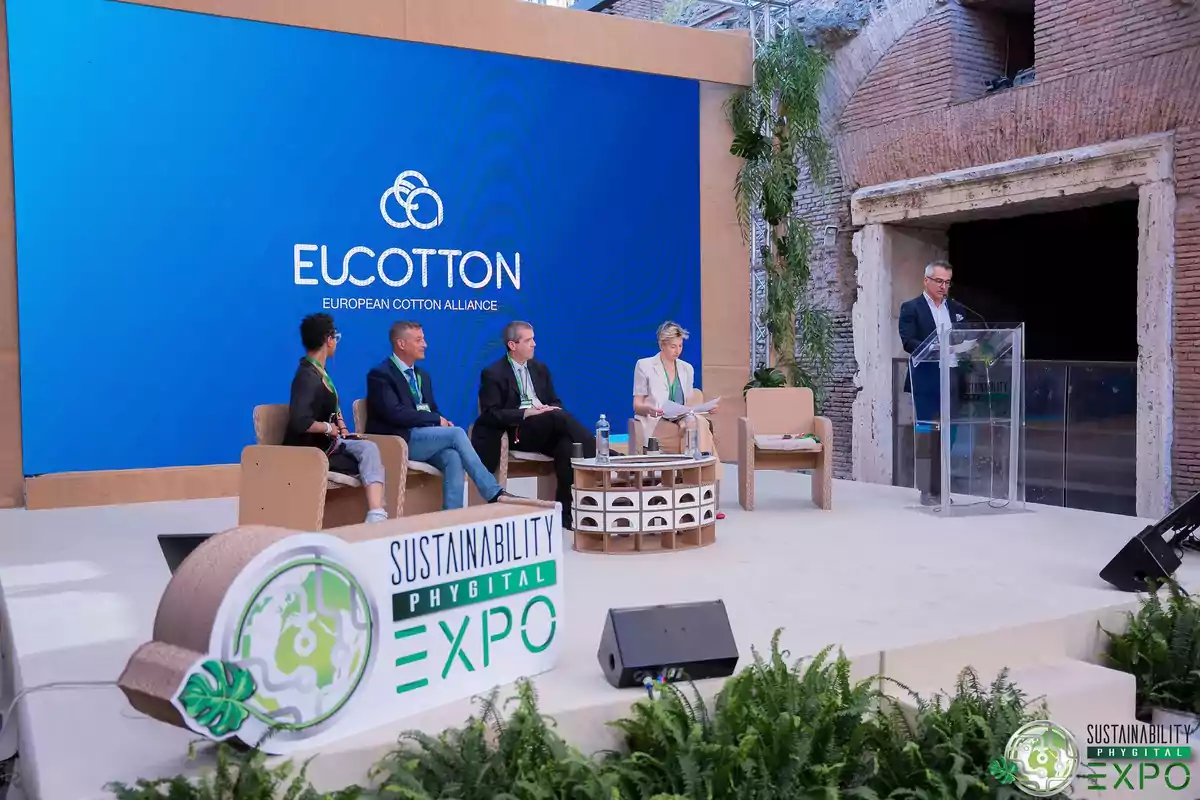 Un grupo de personas participa en una conferencia de la European Cotton Alliance en el Sustainability Phygital Expo, con un orador en un podio y un gran cartel azul de EUCOTTON en el fondo.