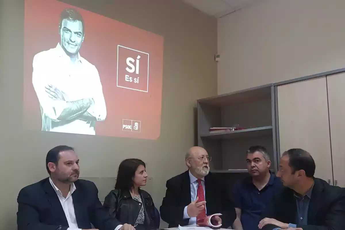 Un grupo de personas sentadas en una mesa discuten mientras en la pared se proyecta una imagen con el texto Un grupo de personas sentadas en una mesa discuten mientras en la pared se proyecta una imagen con el texto