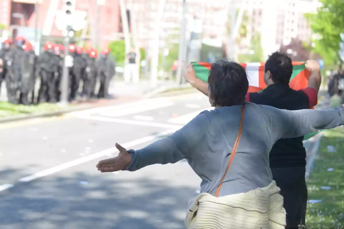 A person with outstretched arms and another holding a flag confront a group of police officers with red helmets on a street.