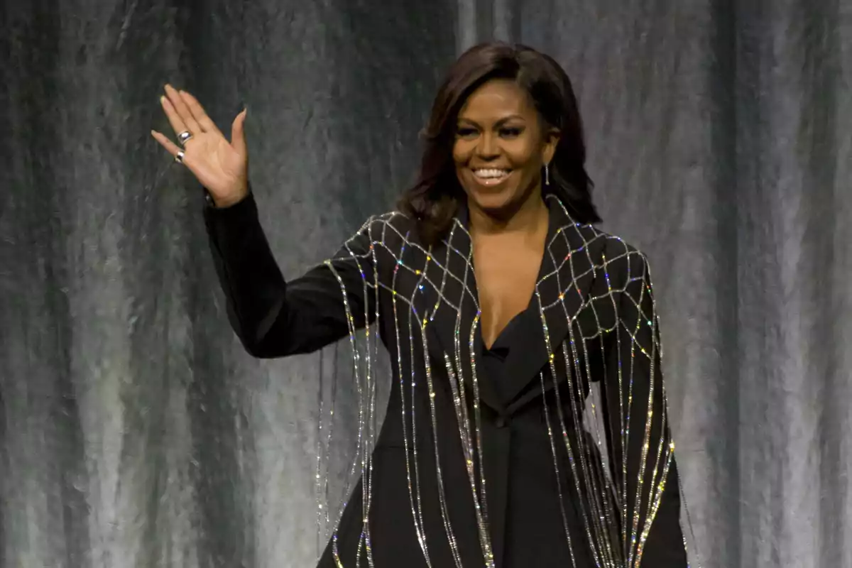 Una persona sonriente con un traje negro decorado con detalles brillantes, levantando la mano en señal de saludo.