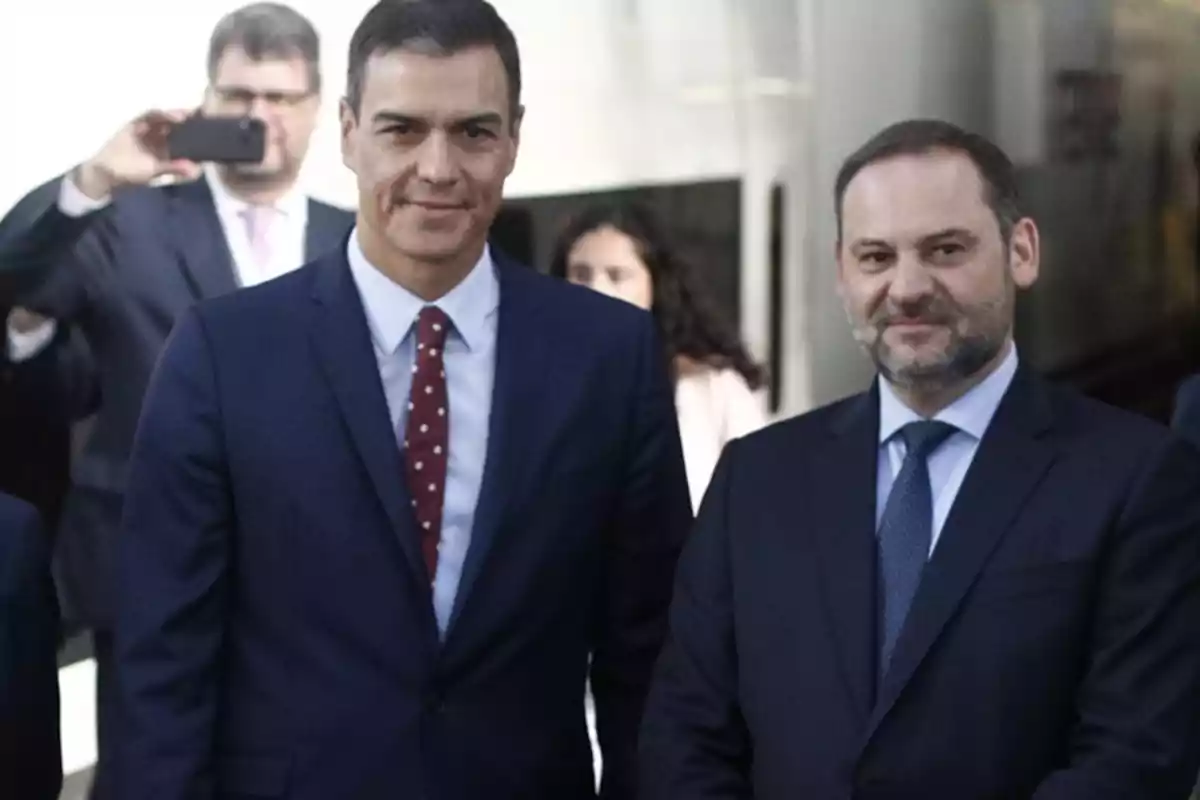 Dos hombres vestidos con trajes oscuros posan para una foto mientras una persona en el fondo toma una foto con su teléfono. Dos hombres vestidos con trajes oscuros posan para una foto mientras una persona en el fondo toma una foto con su teléfono.