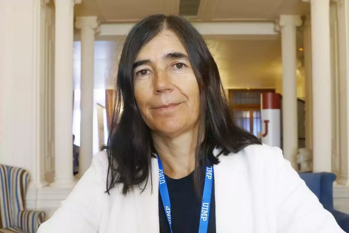 Una persona con cabello oscuro y una chaqueta blanca está sentada en un interior con columnas al fondo.