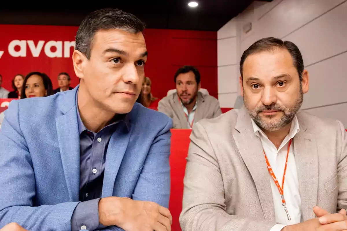 Dos hombres sentados en una sala con fondo rojo y personas al fondo.