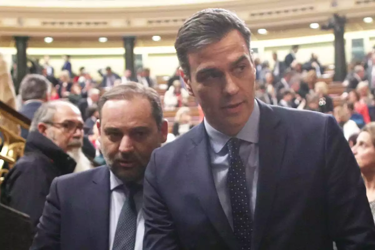 Dos hombres de traje en un entorno formal con personas al fondo.