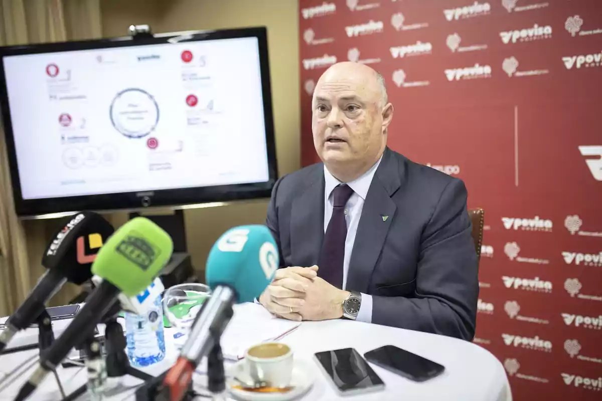 A man in a suit is sitting in front of several microphones at a press conference, with a red background featuring the Povisa logo and a screen behind displaying information.