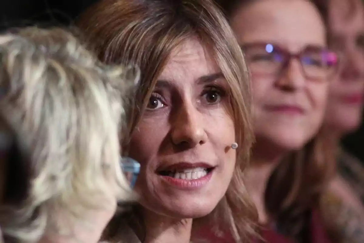 Una mujer hablando con un micrófono en la oreja, con otras personas desenfocadas en el fondo.