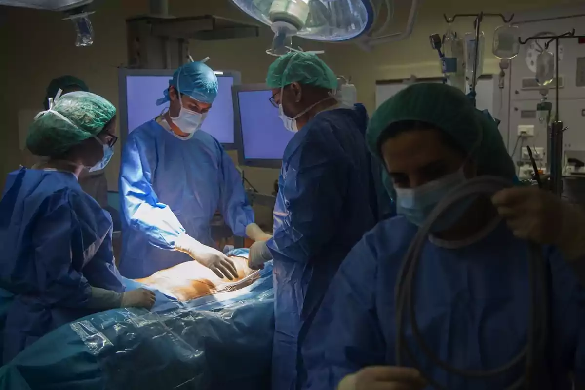 Un grupo de cirujanos realiza una operación en una sala de quirófano, todos vestidos con batas y gorros quirúrgicos azules y mascarillas.