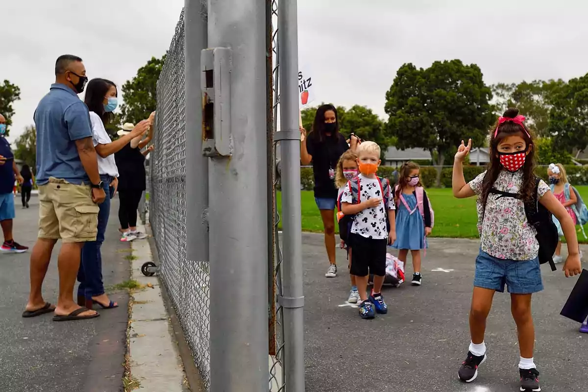 Niños con mascarillas entrando a la escuela mientras los padres los observan desde el otro lado de una cerca.