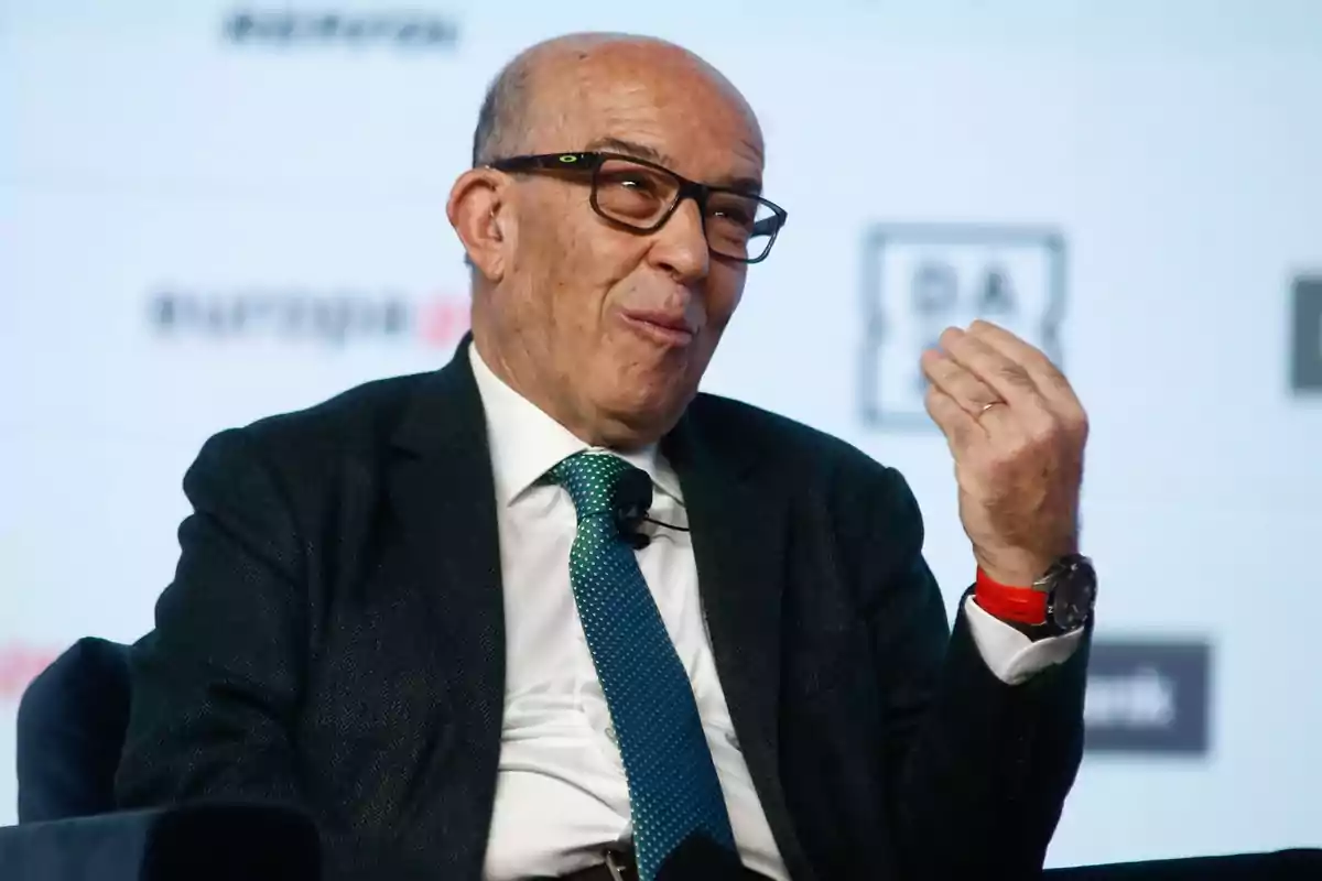 Un hombre calvo con gafas y traje oscuro, corbata verde y reloj rojo, gesticulando con la mano mientras habla en un evento.