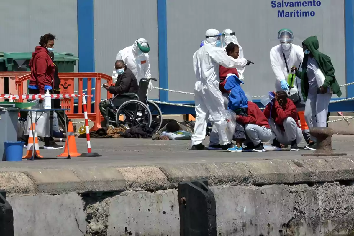 A group of people, some in protective suits, interacts with others on a dock near a Maritime Rescue building.