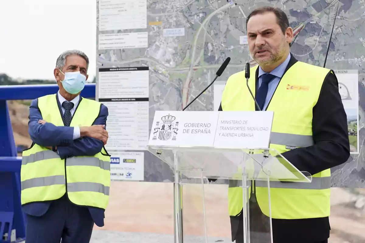 Dos hombres con chalecos reflectantes y traje participan en un evento oficial al aire libre uno de ellos habla en un atril con carteles del Gobierno de España y el Ministerio de Transportes mientras el otro escucha con los brazos cruzados y lleva mascarilla al fondo hay un mapa y documentos técnicos Dos hombres con chalecos reflectantes y traje participan en un evento oficial al aire libre uno de ellos habla en un atril con carteles del Gobierno de España y el Ministerio de Transportes mientras el otro escucha con los brazos cruzados y lleva mascarilla al fondo hay un mapa y documentos técnicos