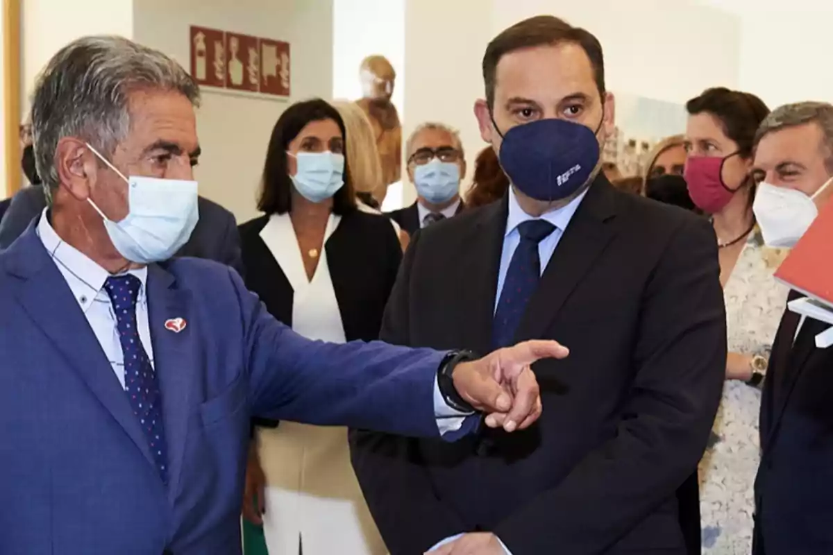 Un grupo de personas con mascarillas en un evento formal mientras un hombre de traje azul señala hacia un lado
