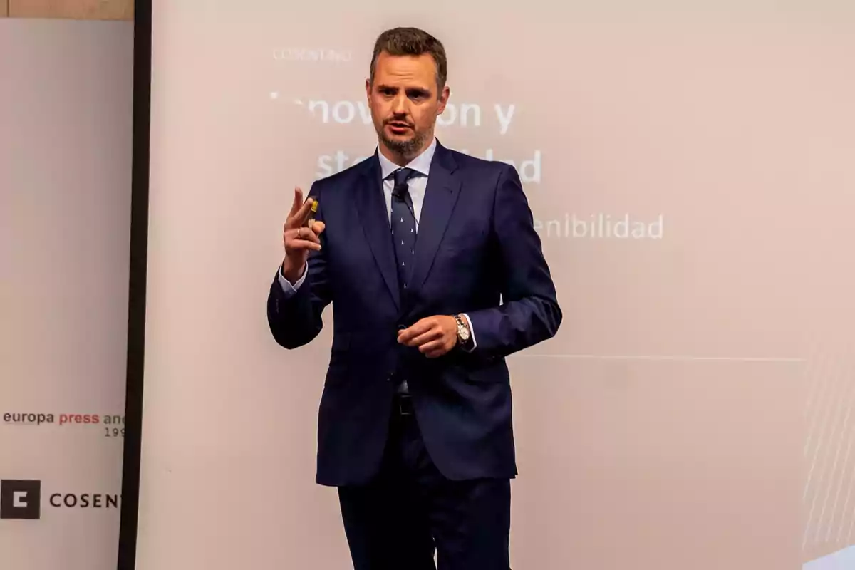 Un hombre en traje azul oscuro está dando una presentación frente a una pantalla con texto parcialmente visible.