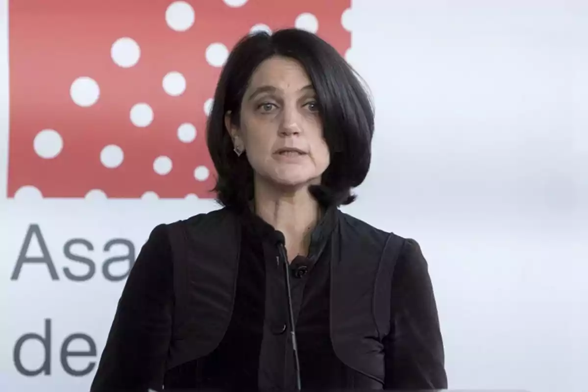 Mujer de cabello oscuro hablando frente a un micrófono con un fondo que tiene un diseño de puntos blancos sobre un fondo rojo.