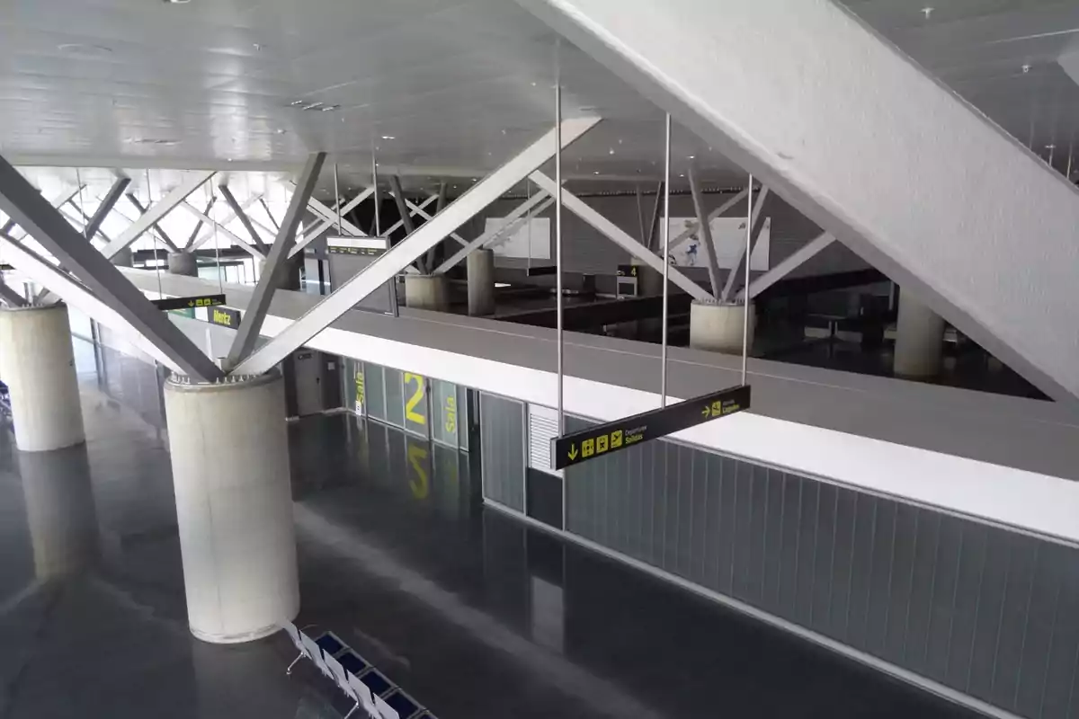 Vista interior de un aeropuerto moderno con estructuras metálicas y columnas de concreto, mostrando señales de dirección y áreas de espera.