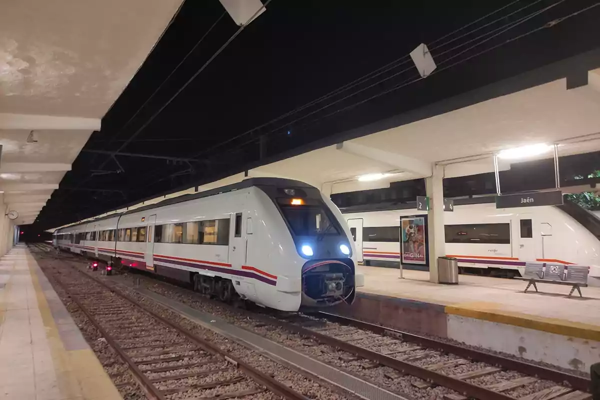 Un tren blanco con franjas rojas y grises está detenido en una estación de tren por la noche con luces encendidas y un cartel que indica el nombre de la estación. Un tren blanco con franjas rojas y grises está detenido en una estación de tren por la noche con luces encendidas y un cartel que indica el nombre de la estación.