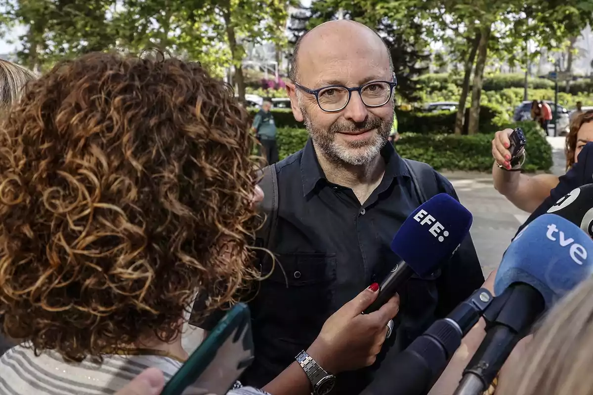 A man with glasses and a beard is being interviewed by several journalists outdoors.