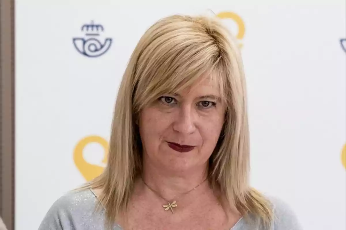 Mujer rubia de cabello lacio y suelto con collar de libélula y fondo blanco con logotipos azules y amarillos. Mujer rubia de cabello lacio y suelto con collar de libélula y fondo blanco con logotipos azules y amarillos.