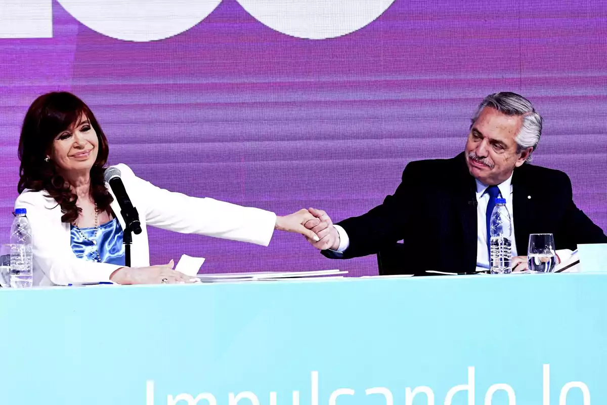 Dos personas se dan la mano en un evento con un fondo púrpura y una mesa azul.