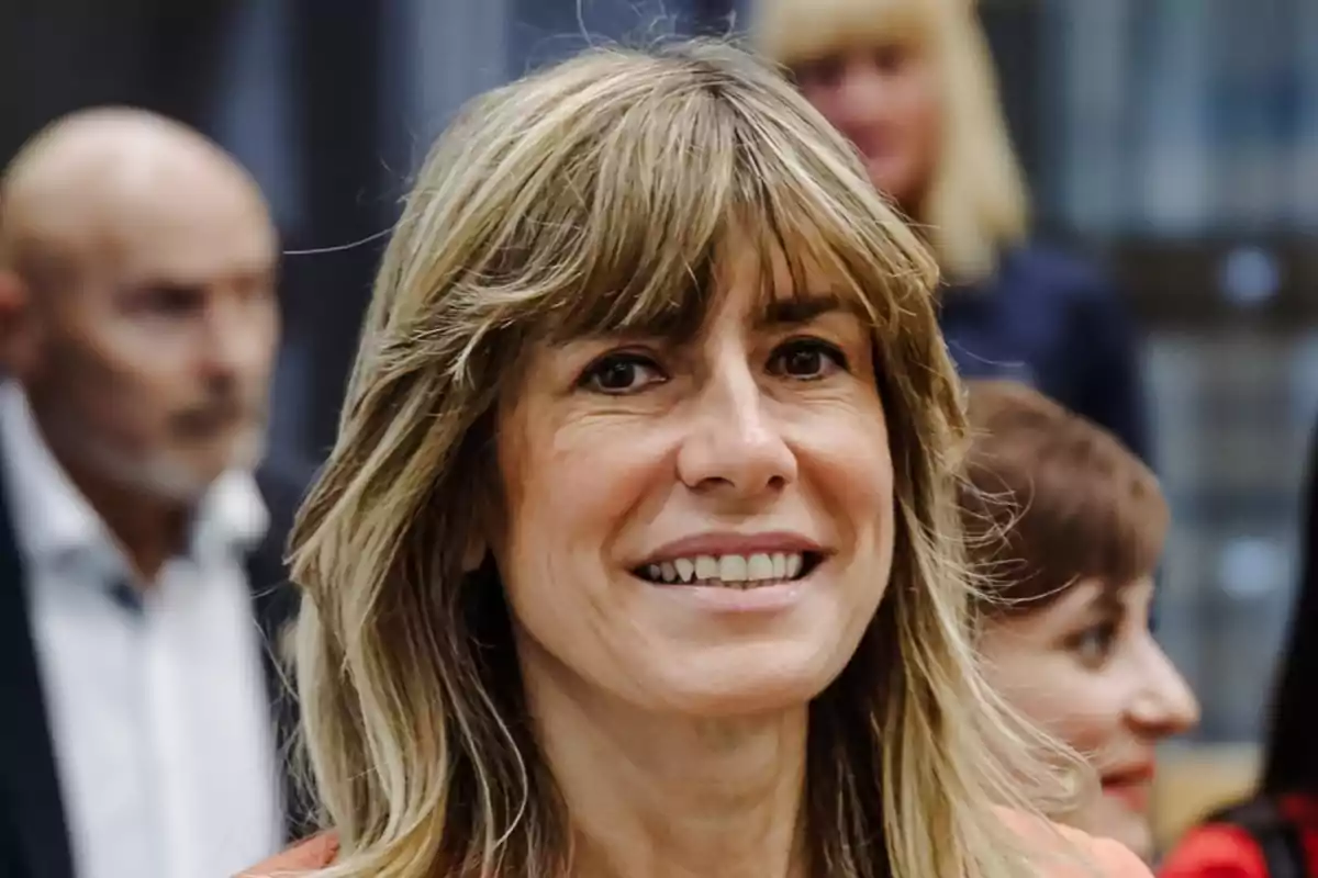 Mujer sonriendo con cabello rubio y flequillo, en un entorno con personas desenfocadas al fondo.