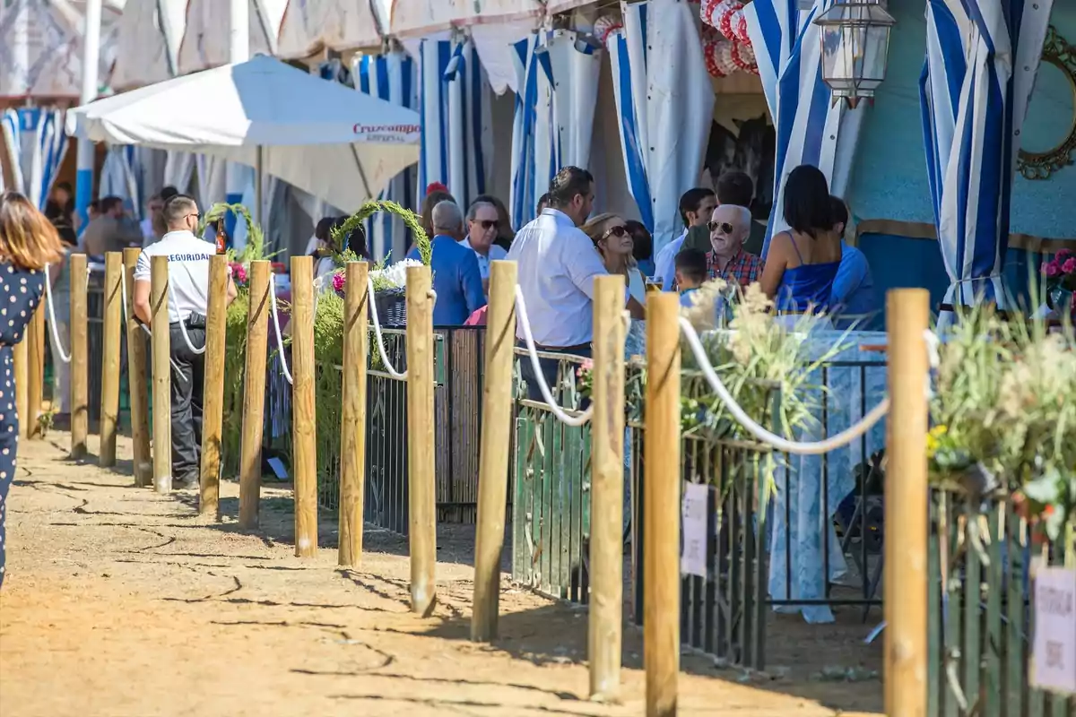 Un grupo de personas disfruta de una feria al aire libre con decoraciones de rayas azules y blancas y un área cercada con postes de madera y cuerdas.