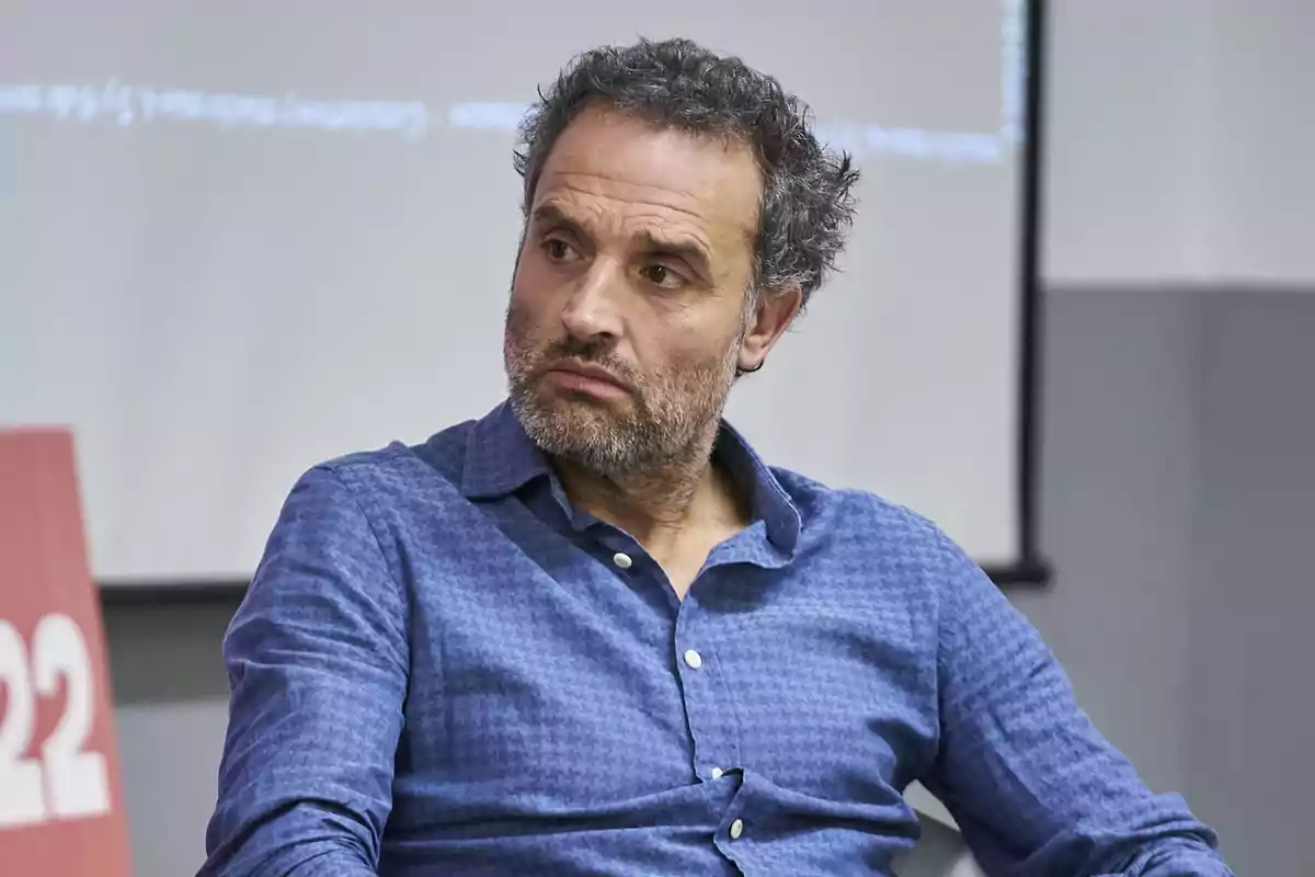 Hombre con barba y cabello rizado, vestido con una camisa azul, sentado y mirando hacia un lado.