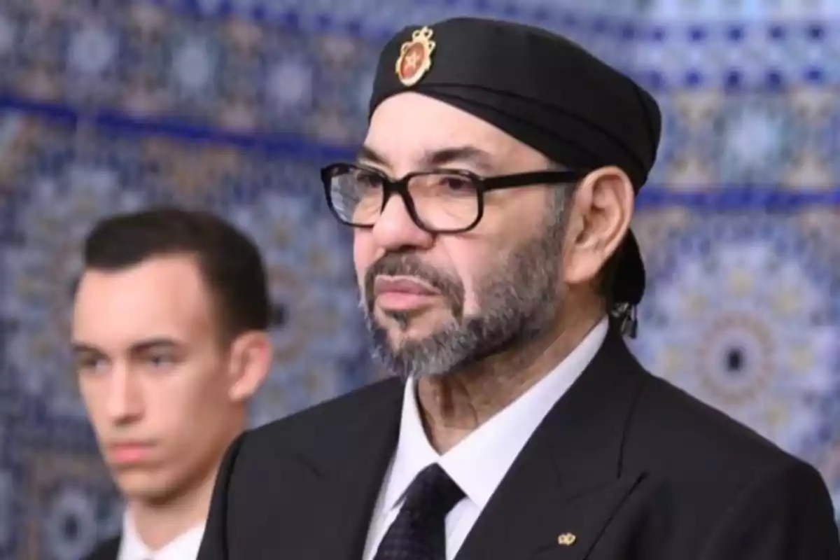 Un hombre con gafas y barba lleva un gorro negro con un emblema dorado en un entorno decorado con azulejos detallados. Un hombre con gafas y barba lleva un gorro negro con un emblema dorado en un entorno decorado con azulejos detallados.