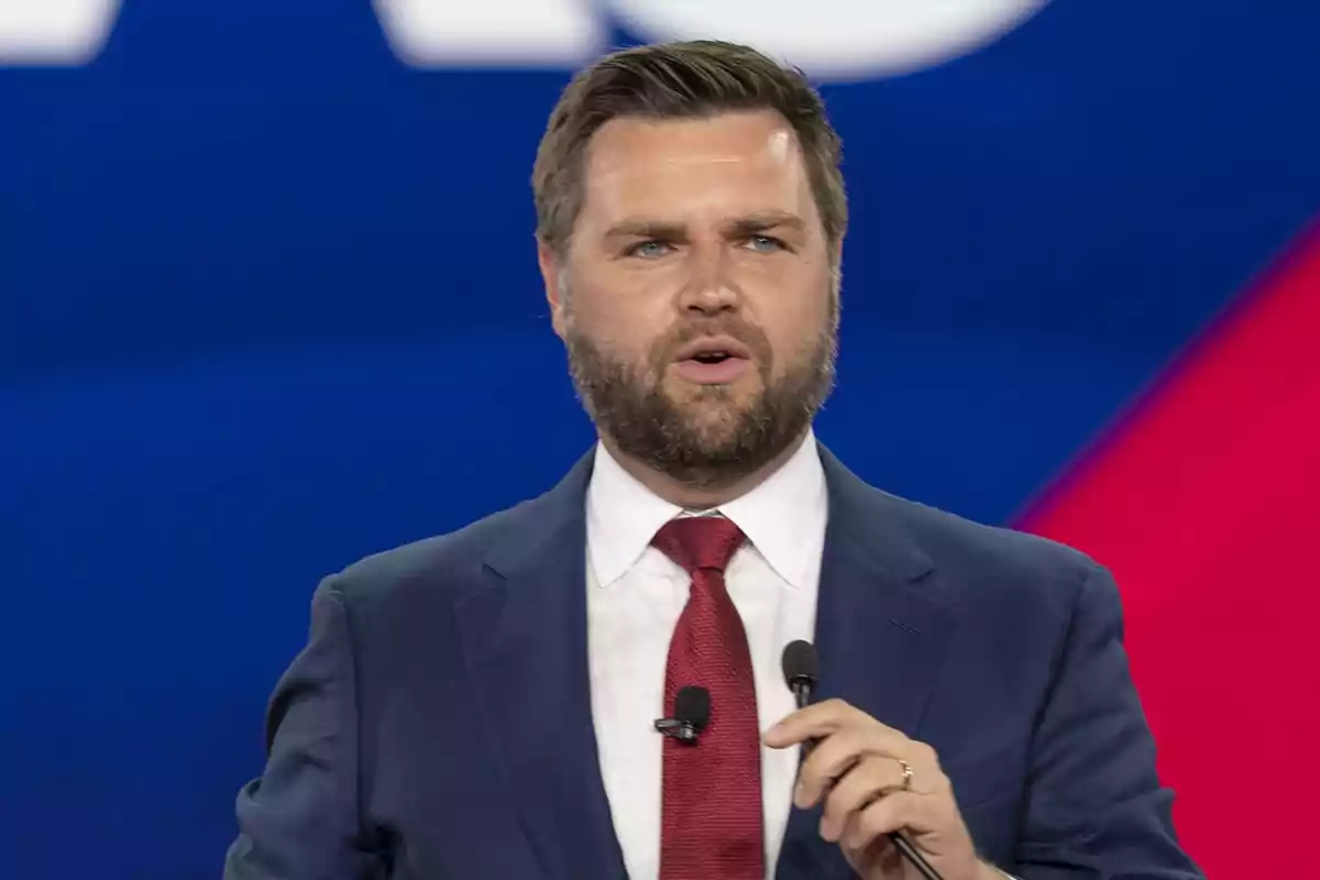 Hombre con barba y traje azul hablando en un evento con fondo azul y rojo.