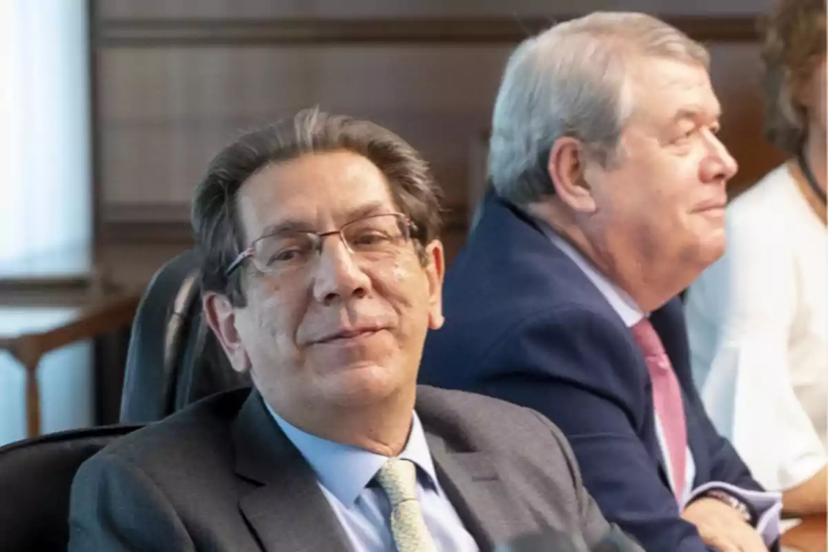 Dos hombres sentados en una reunión, uno de ellos lleva gafas y traje gris, el otro tiene el cabello canoso y viste un traje azul. Dos hombres sentados en una reunión, uno de ellos lleva gafas y traje gris, el otro tiene el cabello canoso y viste un traje azul.