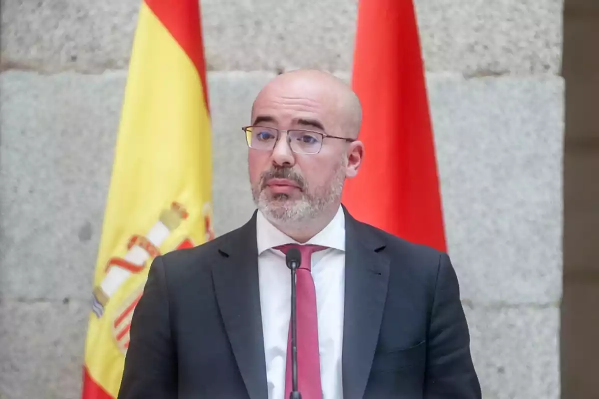 Un hombre con gafas y barba hablando frente a un micrófono con banderas de España de fondo. Un hombre con gafas y barba hablando frente a un micrófono con banderas de España de fondo.