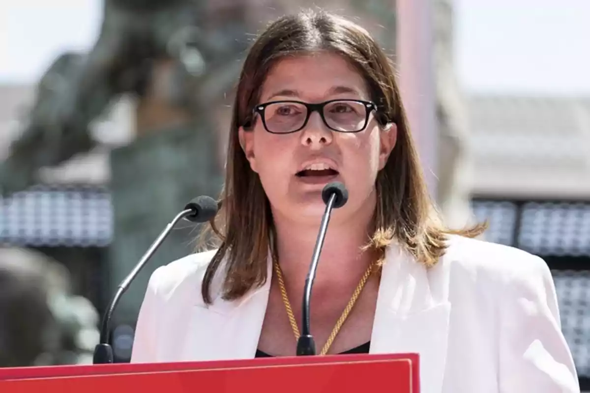 Una persona con gafas y chaqueta blanca hablando en un micrófono al aire libre. Una persona con gafas y chaqueta blanca hablando en un micrófono al aire libre.