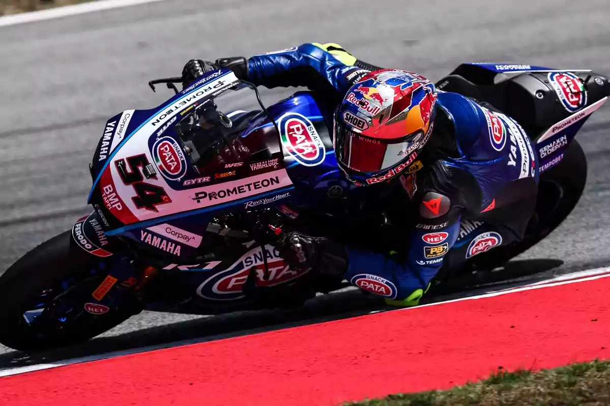 Motociclista en una curva cerrada durante una carrera de motociclismo, vistiendo un traje azul y casco rojo, montando una moto Yamaha con el número 54.