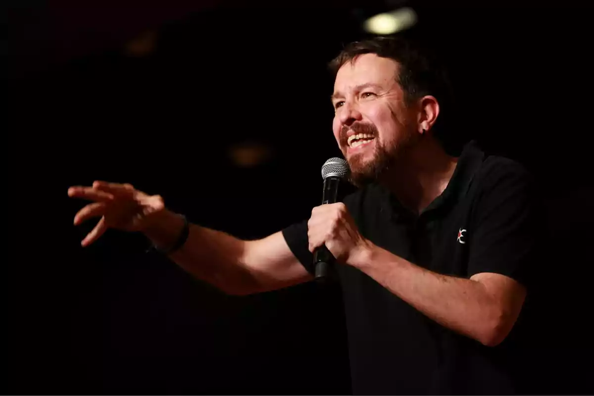 A man with a beard and a black t-jersey holds a microphone while gesturing passionately on a dark stage.