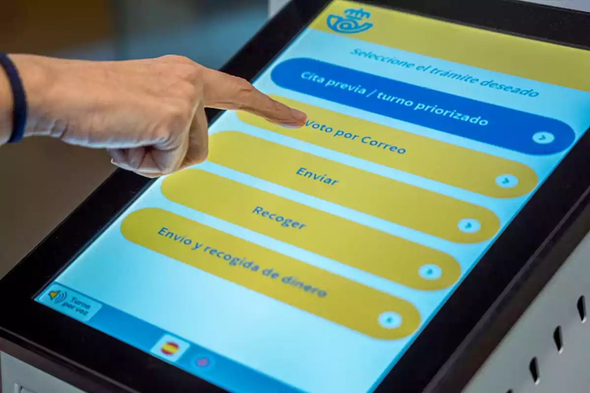 Una mano tocando la pantalla de una máquina de autoservicio en una oficina de correos, seleccionando la opción "Voto por Correo".