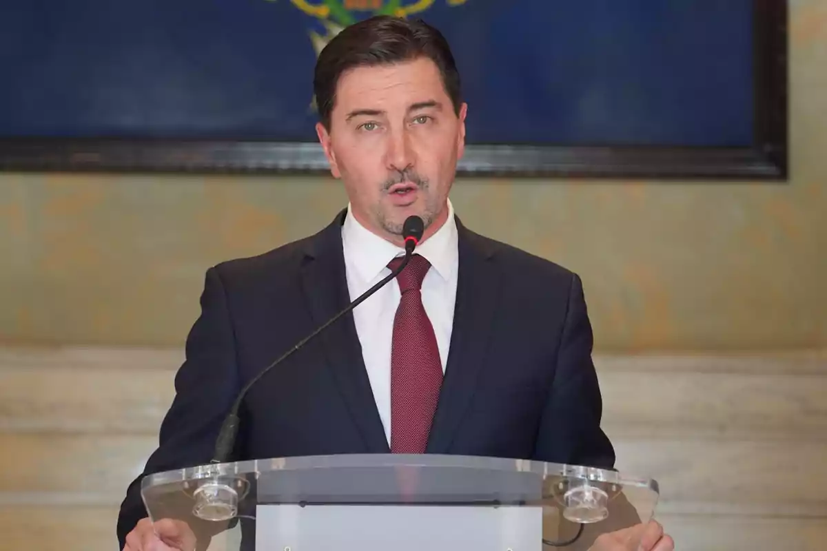 A man in a dark suit and red tie speaks in front of a microphone at a podium.