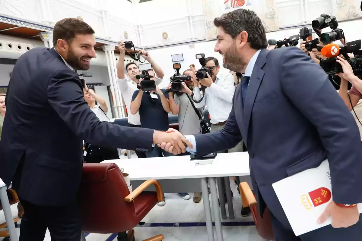 Dos hombres de traje se dan la mano sonrientes mientras varios fotógrafos y camarógrafos capturan el momento en un entorno institucional