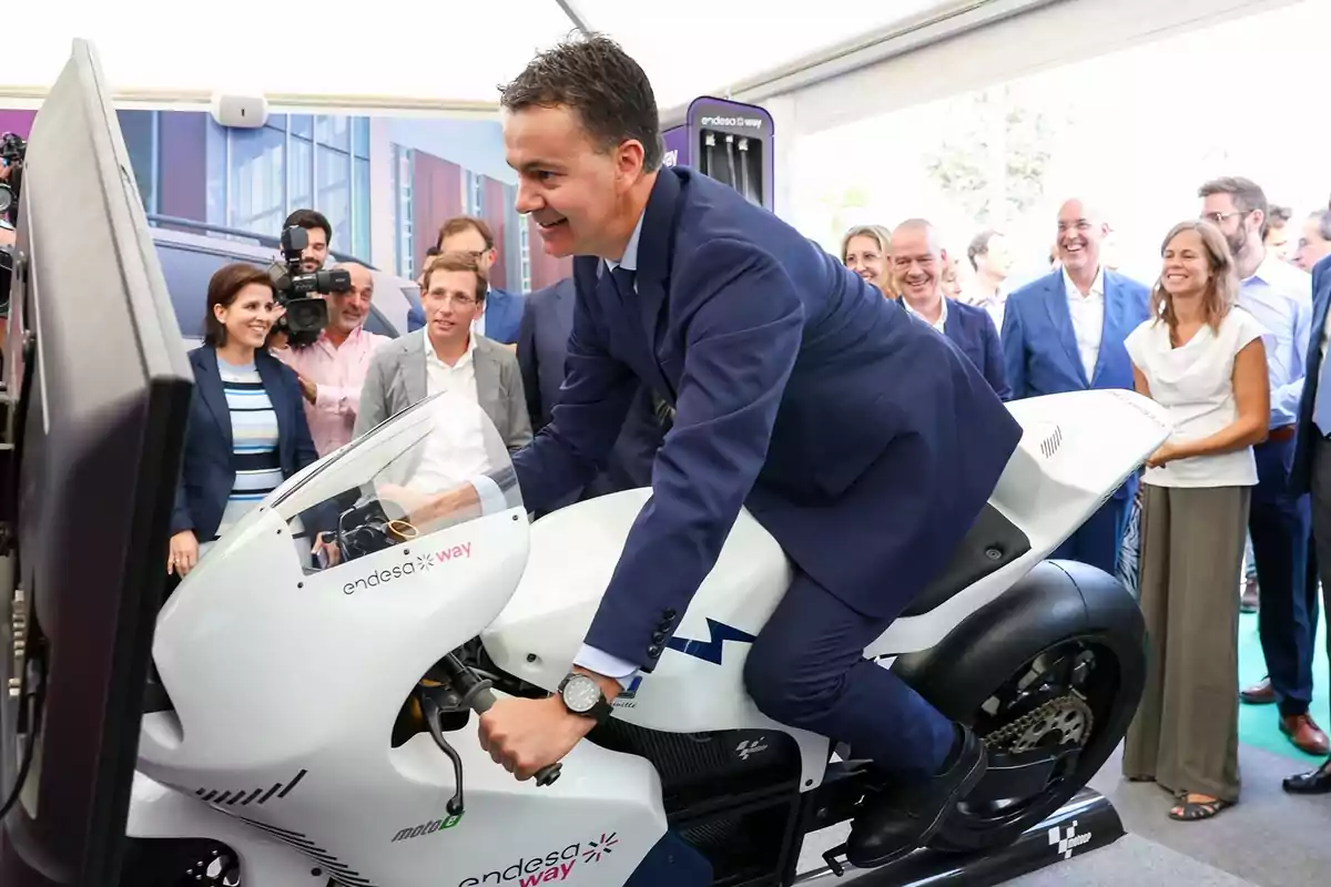 Un hombre en traje monta una motocicleta de exhibición en un evento mientras varias personas observan y sonríen.