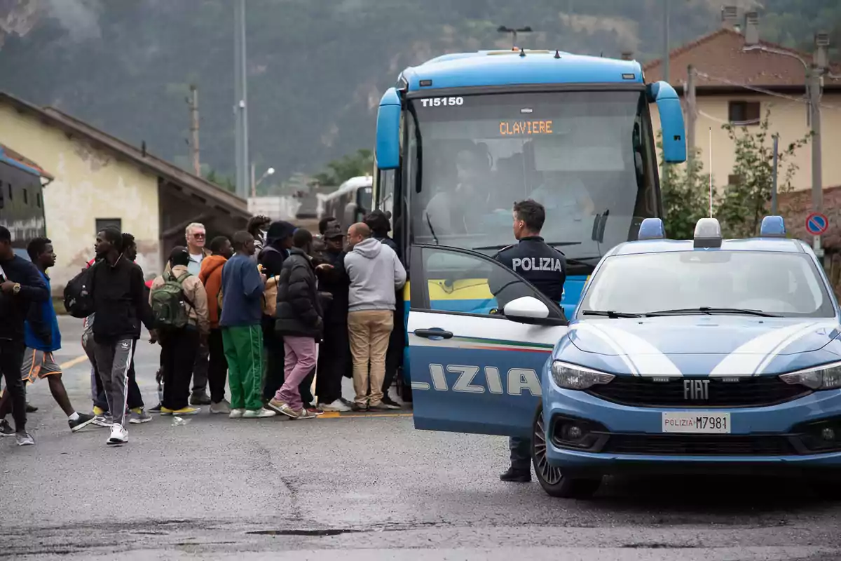 Un grupo de personas se encuentra reunido cerca de un autobús con la palabra "Claviere" en el letrero, mientras un oficial de policía está de pie junto a un coche patrulla en una zona urbana.