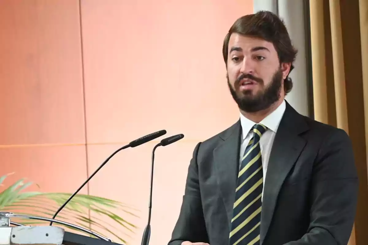 Hombre con barba y traje oscuro hablando en un podio con micrófonos.