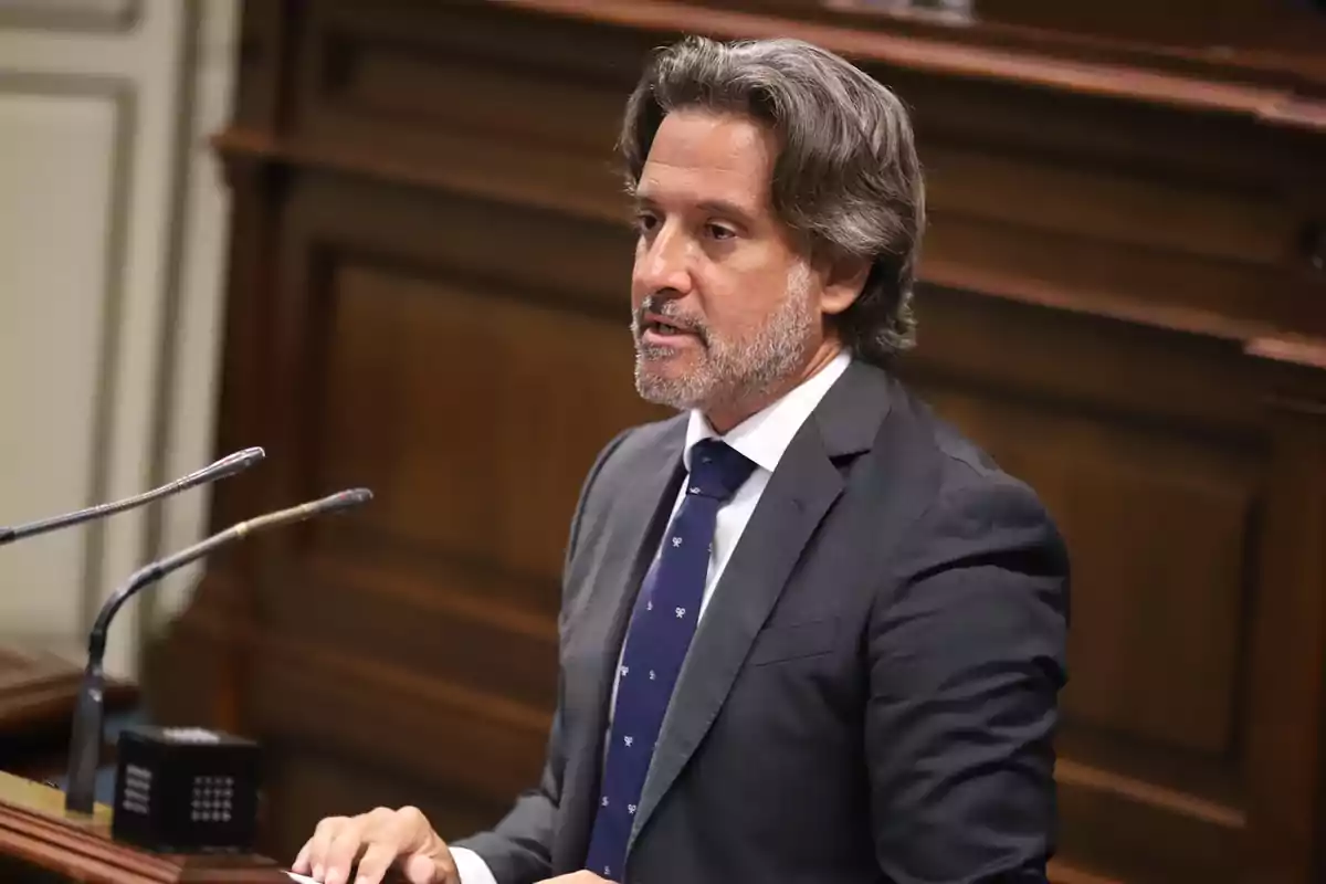 Un hombre de traje y corbata habla en un podio con micrófonos en un entorno formal. Un hombre de traje y corbata habla en un podio con micrófonos en un entorno formal.