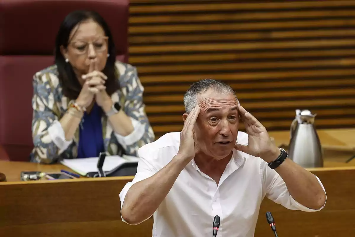 A man gestures with his hands on his head while speaking in a formal setting with a woman sitting behind watching.