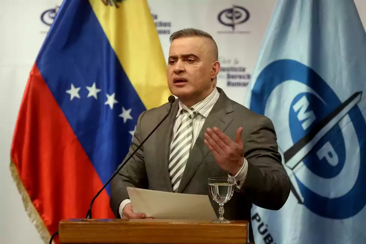 Hombre en traje gris hablando en un podio con banderas de Venezuela y otra organización de fondo.