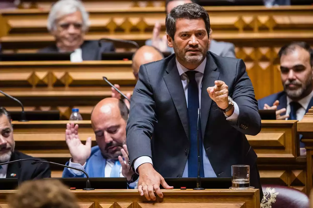 Un hombre de traje habla en un parlamento mientras otros lo observan y aplauden. Un hombre de traje habla en un parlamento mientras otros lo observan y aplauden.