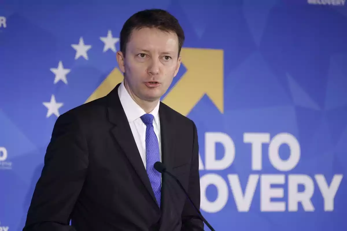 Hombre de traje y corbata azul hablando frente a un micrófono con fondo azul y amarillo que tiene estrellas blancas y un texto parcialmente visible Hombre de traje y corbata azul hablando frente a un micrófono con fondo azul y amarillo que tiene estrellas blancas y un texto parcialmente visible