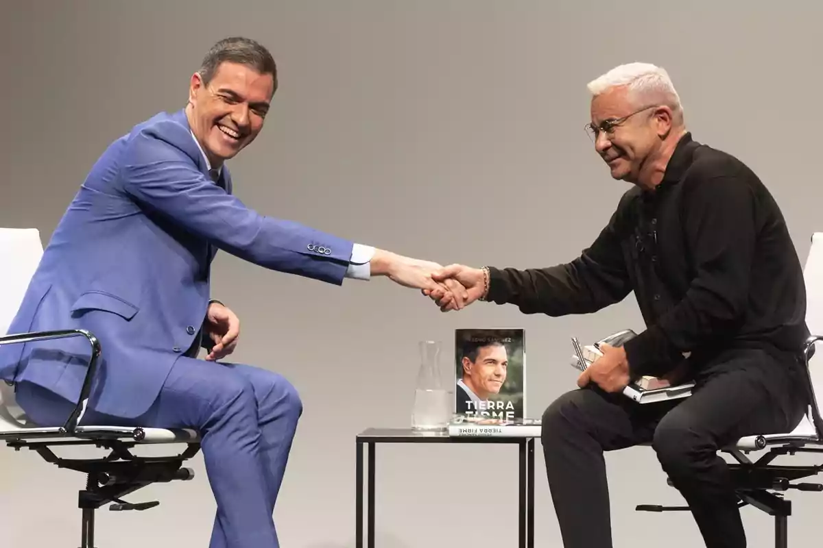 Dos hombres se dan la mano mientras están sentados en sillas, uno de ellos sostiene un libro titulado "Tierra Firme" sobre una mesa.